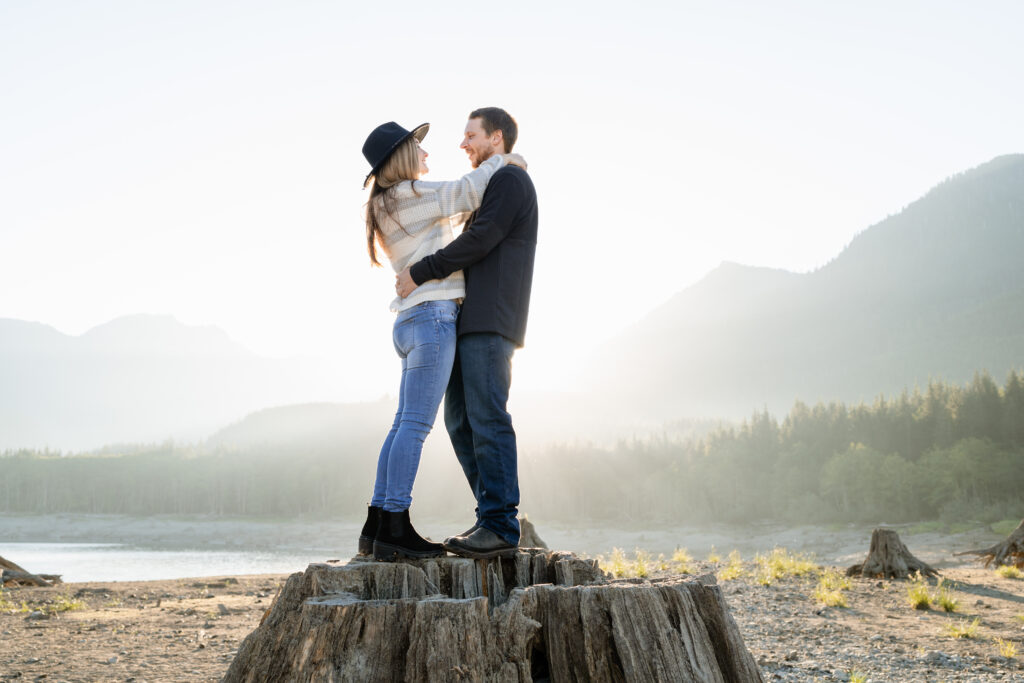 A couple embracing and smiling at each other while standing on a stump with the Cascade mountains of Washington behind them. The sun is rising over the mountains that they stand in front of.