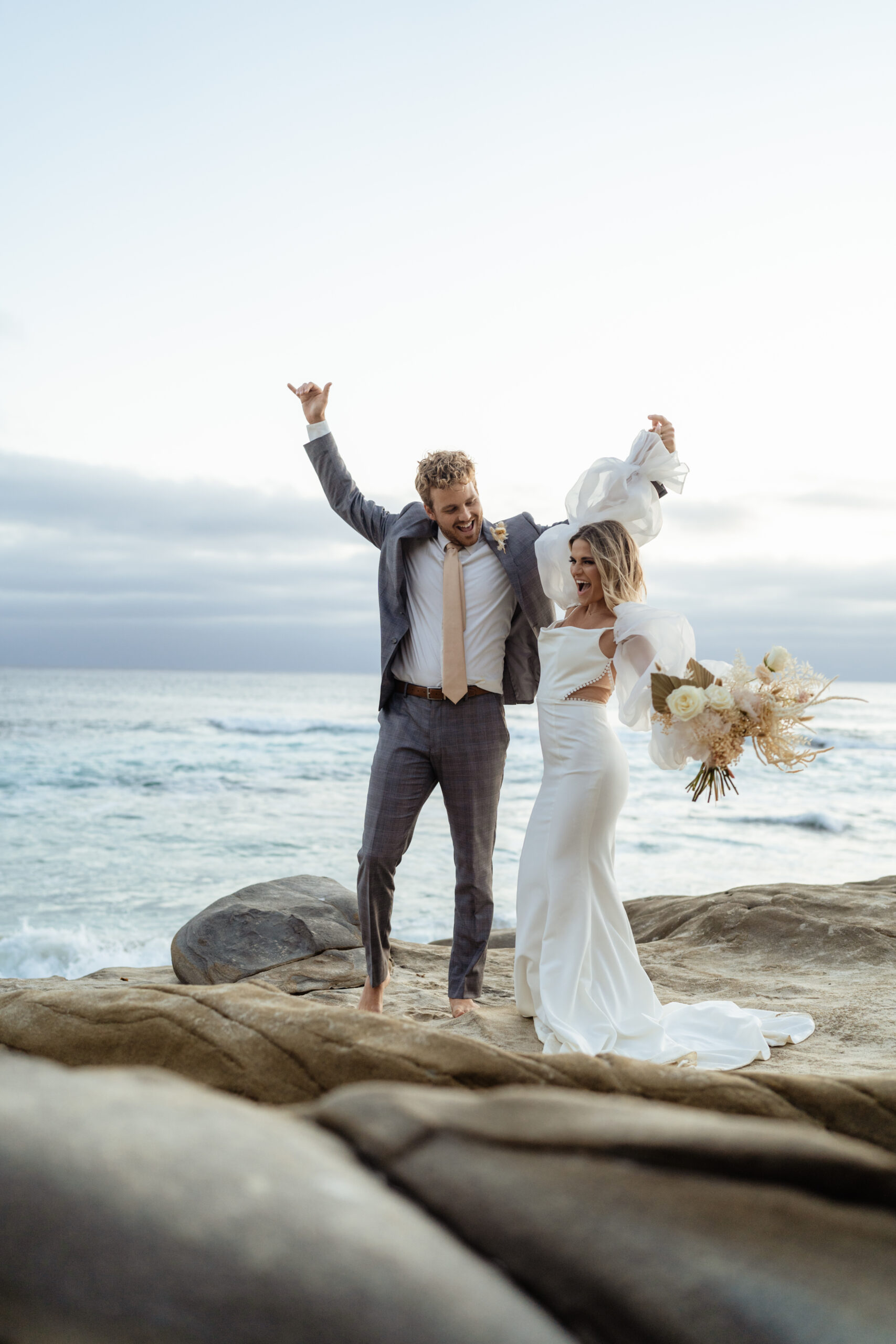 A couple in wedding attire cheering with their hands help up right after eloping on the beach. They are standing on a rock with the ocean behind them. The bride is holding a bouquet of flowers.