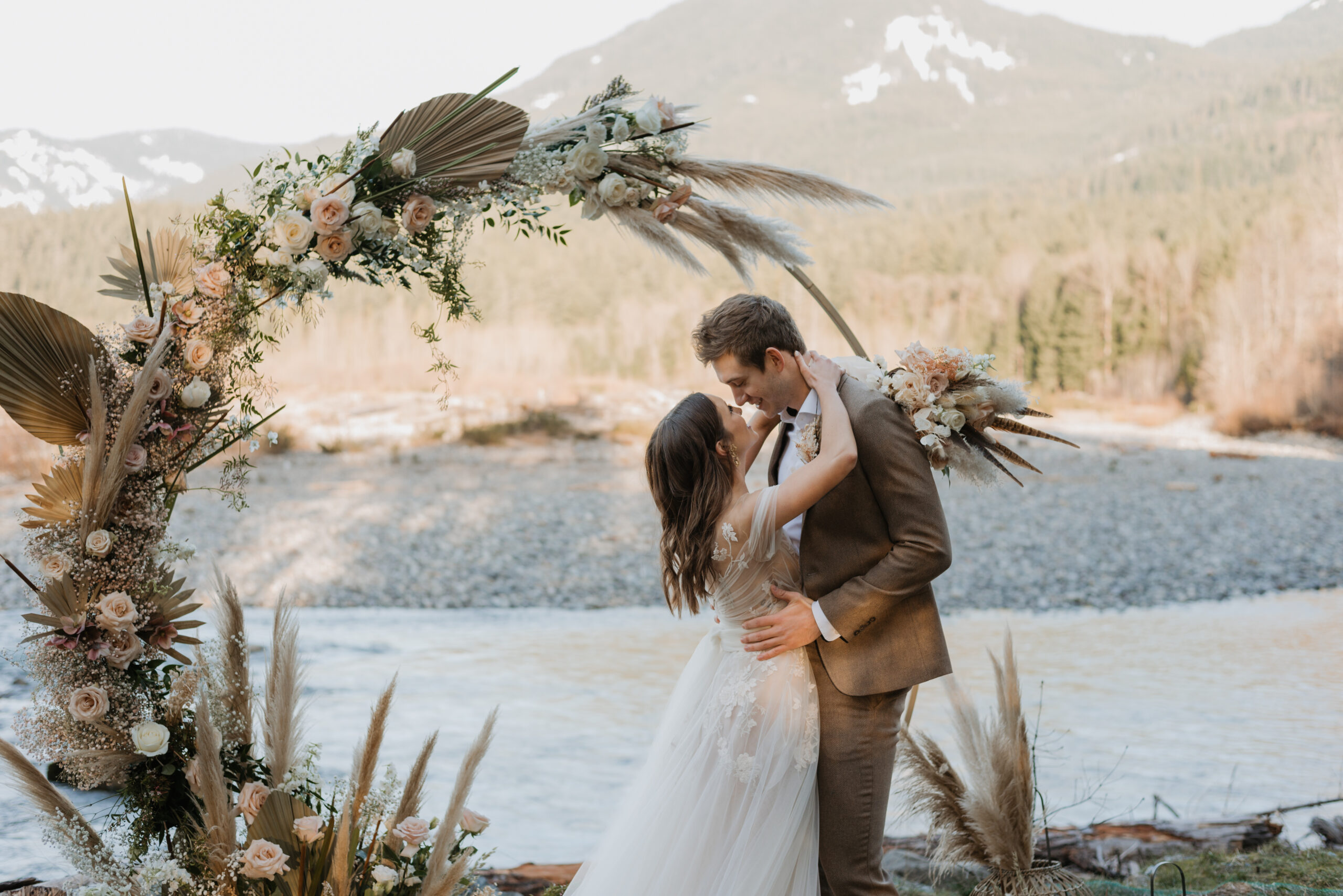A couple eloping in Washington State. They are wearing wedding attire and standing in front of a river and mountain view. There is a boho floral arch behind them. They are embracing each other and going in for a kiss.