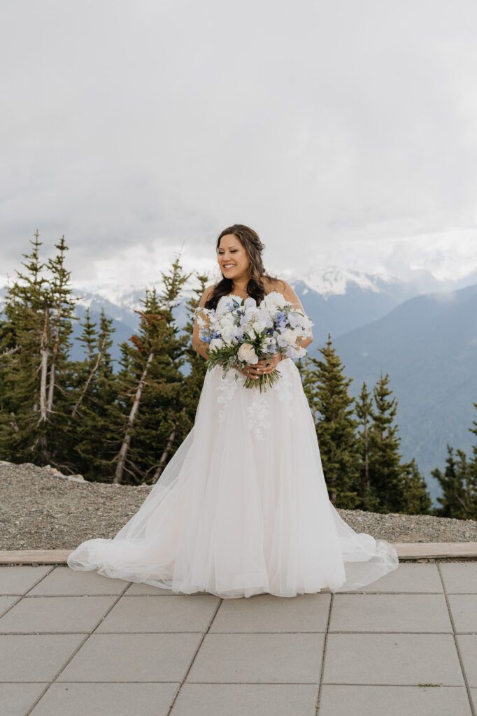 A bride holds her bouquet and smiles while standing atop Mt. Rainer Platform with the trees and mountains of Washington state in the background. 