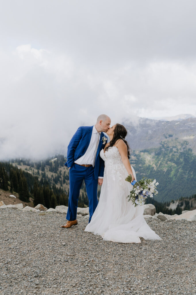 A bride and groom share a kiss on top of Crystal Mountain in Washington State. The mountains and clouds are in the background. 