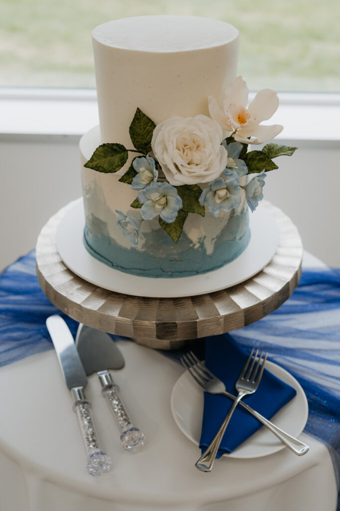 A wedding cake with white and blue frosting. There are white and blue flowers between the first and second tier.