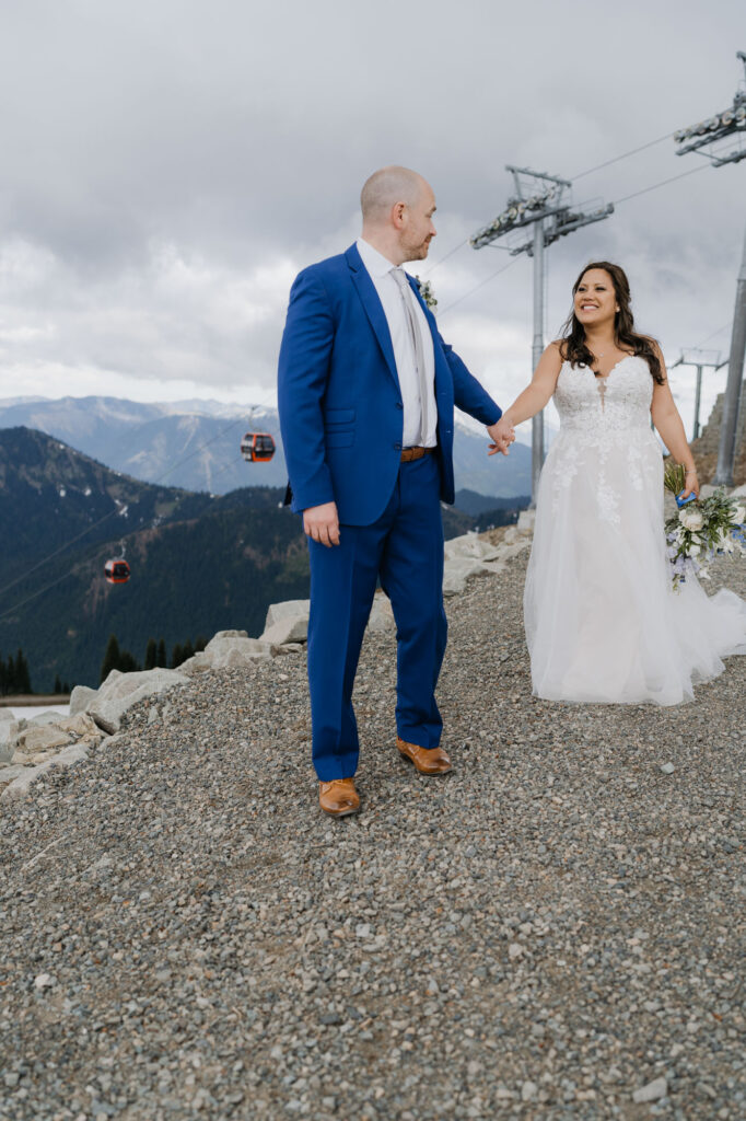 A bride and groom hold hands while walking atop Crystal mountain with a view of the gondola in the background.