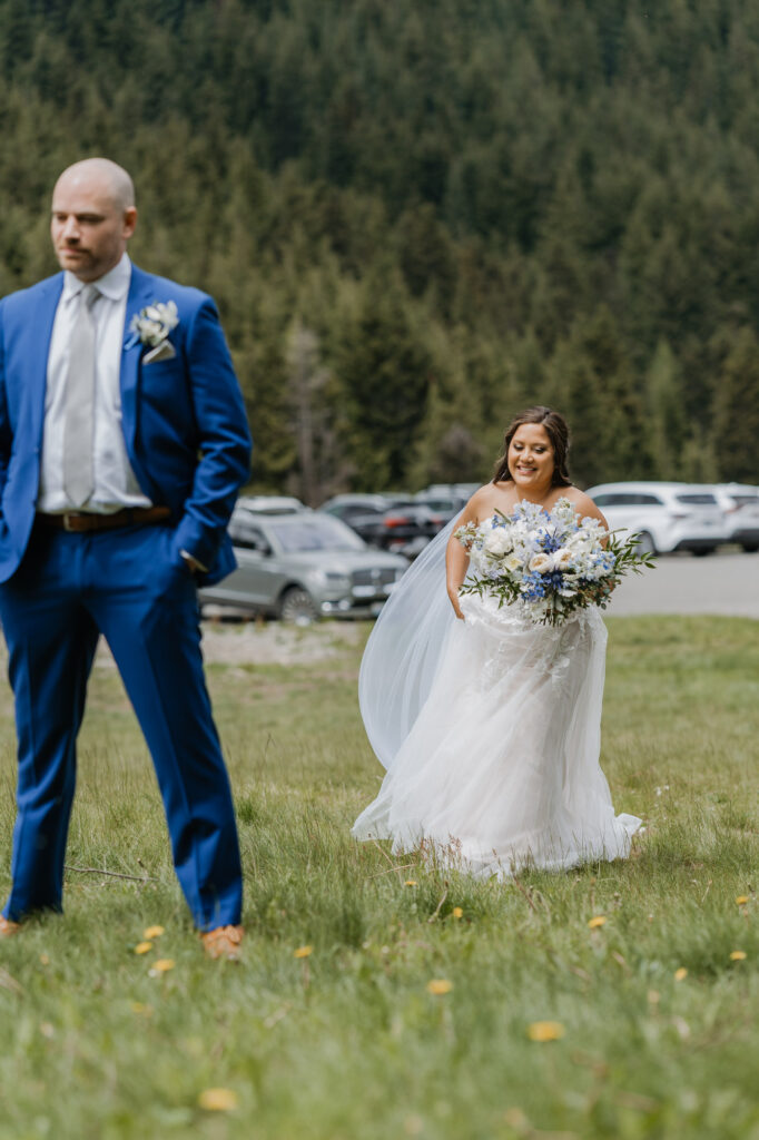 A bride walks toward the groom for their first look while his back is facing her at the base of Crystal Mountain Resort. 