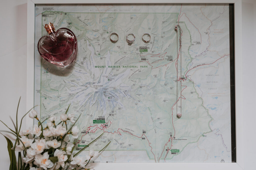 Wedding day details on top of a map of Mt. Rainier National Park. There is a bottle of perfume, 3 rings, a necklace, and some flowers.