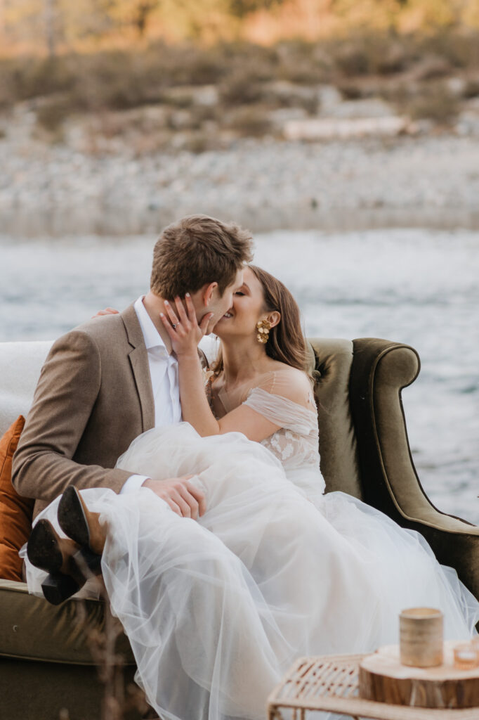 A bride and groom kissing while sitting on a small couch in front of a river in Washington