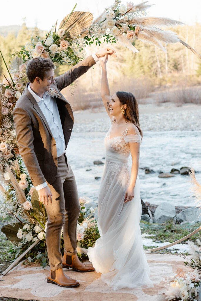 A couple smiling at each other while twirling under a flower arch at their Washington elopement along the river
