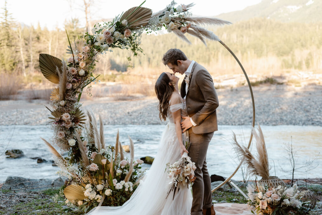A couple having a Washington elopement in front of a flower arch along the river.