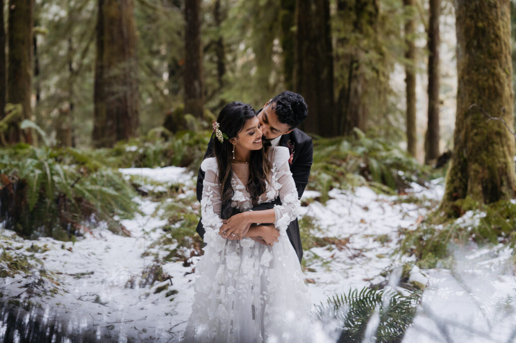 A Washington forest elopement. The couple is looking down while embracing each other in the forest. There is snow on the ground and moss on the trees.